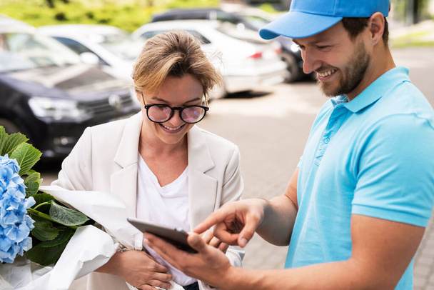 Young and professional delivery man in blue uniform delivers flowers to a woman client - Foto, Bild