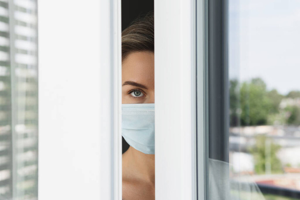 Молодая напуганная женщина в защитной маске смотрит в окно - Фото, изображение