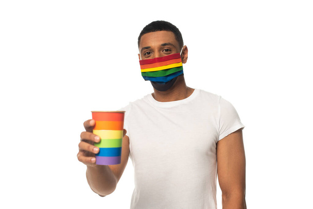 hombre afroamericano con máscara de seguridad y taza de papel en colores arco iris aislado en blanco, concepto lgbt - Foto, imagen
