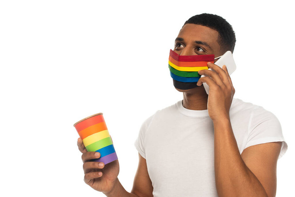 Αφροαμερικάνος άνδρας με ιατρική μάσκα και χάρτινο κύπελλο σε χρώματα lgbt μιλώντας στο κινητό τηλέφωνο απομονωμένο σε λευκό - Φωτογραφία, εικόνα