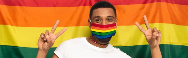 african american man in rainbow colors medical mask showing victory gesture on background of lgbt flag, banner - Photo, Image