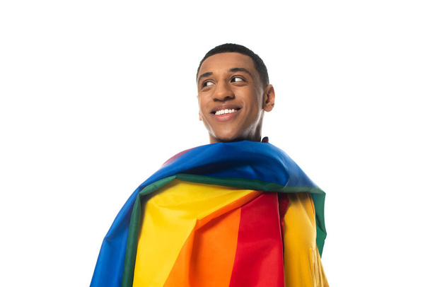 hombre afroamericano feliz, cubierto con la bandera del lgbt, mirando lejos aislado en blanco - Foto, Imagen