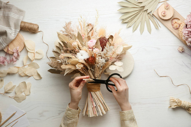 Florist making beautiful bouquet of dried flowers at white table, top view - 写真・画像