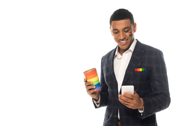 Африканский американский бизнесмен с покрашенным на лице флагом lgbt держит кофе, чтобы пойти и смартфон изолирован на белом - Фото, изображение