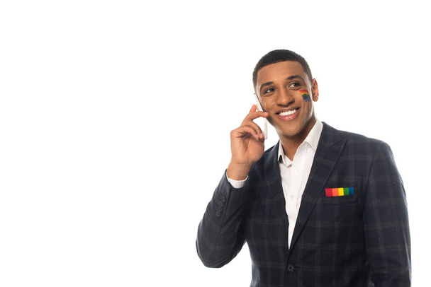 alegre hombre de negocios afroamericano con bandera lgbt pintado en la cara hablando en el teléfono móvil aislado en blanco - Foto, Imagen