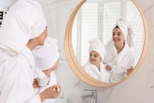 Young mother applying mask onto daughter's face near mirror in bathroom - Φωτογραφία, εικόνα