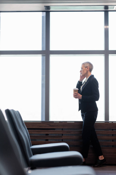 Πλευρική άποψη της ώριμης επιχειρηματία με καφέ για να πάει μιλάμε στο κινητό τηλέφωνο κοντά στο παράθυρο  - Φωτογραφία, εικόνα