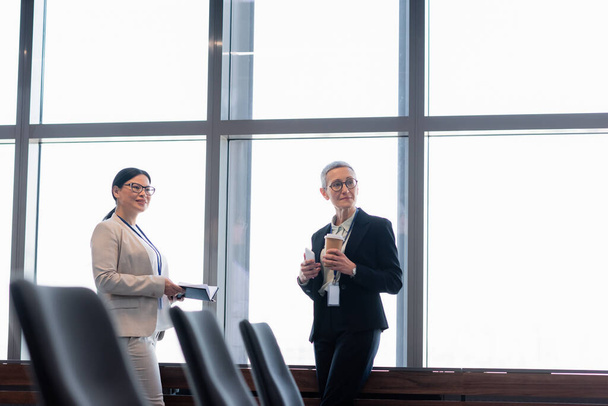 Des femmes d'affaires interraciales souriantes avec du café à emporter et un cahier détournant les yeux au bureau  - Photo, image