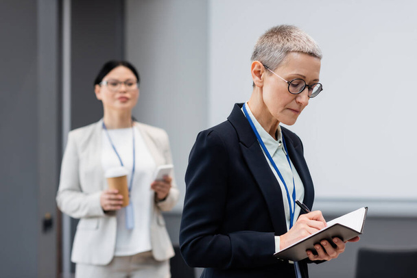 Mature businesswoman writing on notebook near asian colleague on blurred background  - Photo, Image
