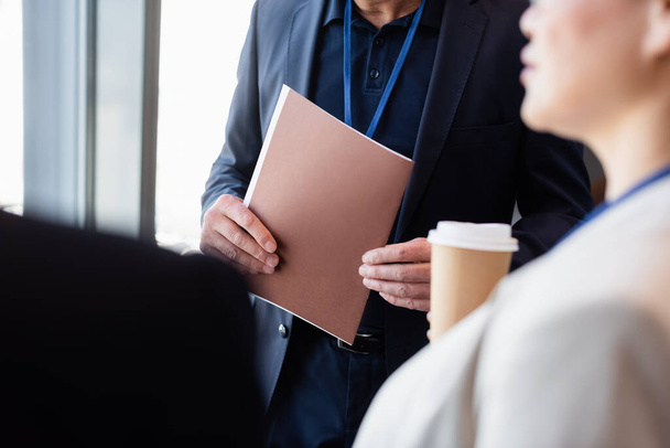 Cropped view of businessman holding paper folder near colleague with coffee to go on blurred foreground  - Фото, изображение