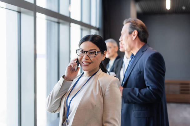 Азиатская бизнесвумен разговаривает на смартфоне рядом с размытыми коллегами  - Фото, изображение