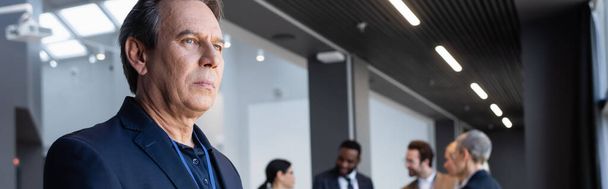 Mature businessman looking away near blurred business people in office, banner  - Foto, afbeelding