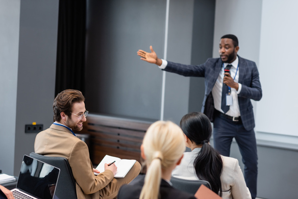 Homme d'affaires souriant avec un cahier regardant un collègue près d'un orateur afro-américain flou dans la salle de conférence  - Photo, image