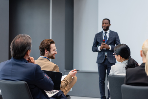 Un sonriente hombre de negocios hablando con un colega casi borroso afroamericano en la sala de conferencias  - Foto, Imagen