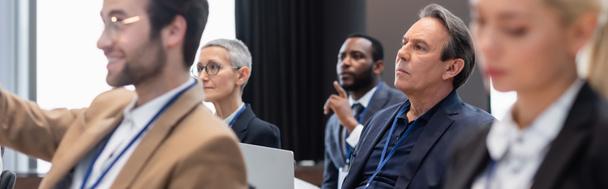 Empresário de meia-idade com laptop sentado perto de colegas multiétnicos durante o seminário, banner  - Foto, Imagem