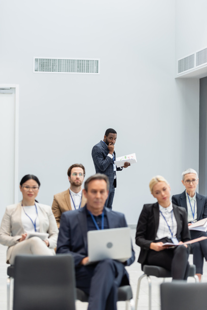 Hombre de negocios afroamericano sosteniendo documentos cerca de gente de negocios interracial borrosa en sala de conferencias  - Foto, imagen