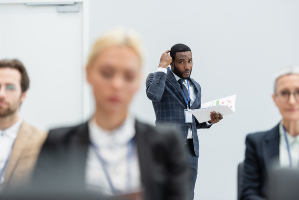 Homme d'affaires afro-américain pensif tenant des papiers près de collègues flous pendant la formation   - Photo, image