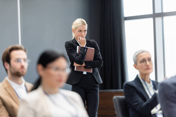 Donna d'affari concentrata con cartellina di carta in piedi vicino a colleghi sfocati in sala conferenze  - Foto, immagini