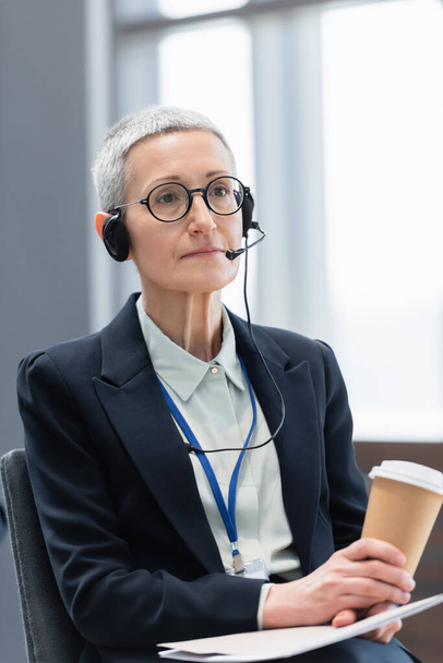 Mature businesswoman in headset holding papers and coffee to go during conference  - Photo, Image