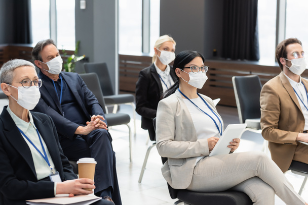 医療用マスクを着た異人種間のビジネス関係者がコーヒーを飲みながらセミナーに座り - 写真・画像