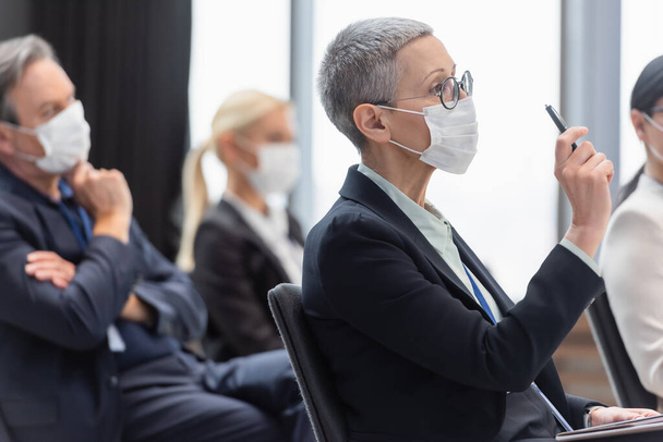 ぼやけた同僚の近くでセミナー中にペンを持っている医療用マスクの成熟したビジネスウーマンの選択的な焦点 - 写真・画像