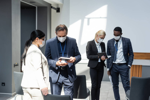 gente de negocios interracial con máscaras médicas sosteniendo cuadernos mientras habla durante el descanso en el seminario - Foto, Imagen