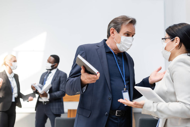 Reifer Geschäftsmann in medizinischer Maske im Gespräch mit asiatischen Kollegen während der Konferenz - Foto, Bild