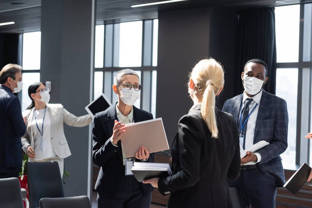 maduro mujer de negocios en médico máscara celebración de documentos mientras habla con interracial colegas - Foto, imagen