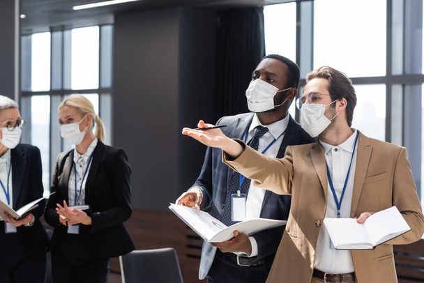 young businessman in medical mask pointing with hand during conversation with african american colleague - Photo, Image