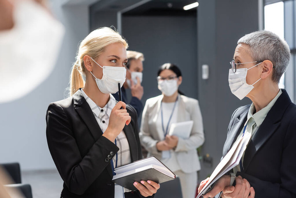 młoda bizneswoman w masce medycznej trzymając długopis i notatnik w pobliżu dojrzałego kolegi - Zdjęcie, obraz