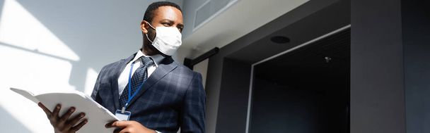 african american businessman in medical mask holding documents while looking away in office, banner - Foto, immagini