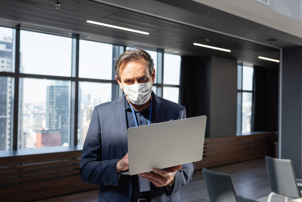 middle aged businessman in medical mask using laptop while standing in office - Fotó, kép