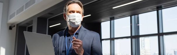 businessman in protective mask looking away while standing with laptop in office, banner - Foto, Bild