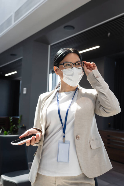 азіатська бізнес-леді тримає мобільний телефон і коригує окуляри, дивлячись в офіс
 - Фото, зображення