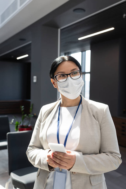 azjatycki bizneswoman w medyczny maska i okulary gospodarstwa smartfon w biuro - Zdjęcie, obraz