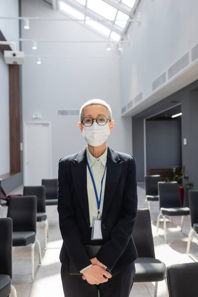 donna d'affari di mezza età in maschera medica guardando la fotocamera in sala conferenze - Foto, immagini