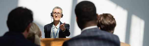спикер среднего возраста, указывая на размытых бизнесменов во время семинара, баннер - Фото, изображение