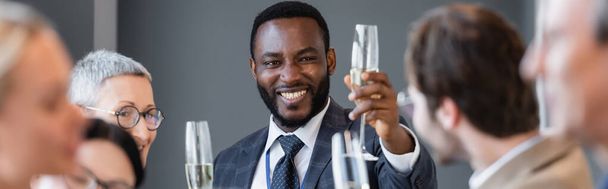 uomo d'affari afro-americano sorridente che tiene bicchiere di champagne vicino al collega in primo piano sfocato, striscione - Foto, immagini
