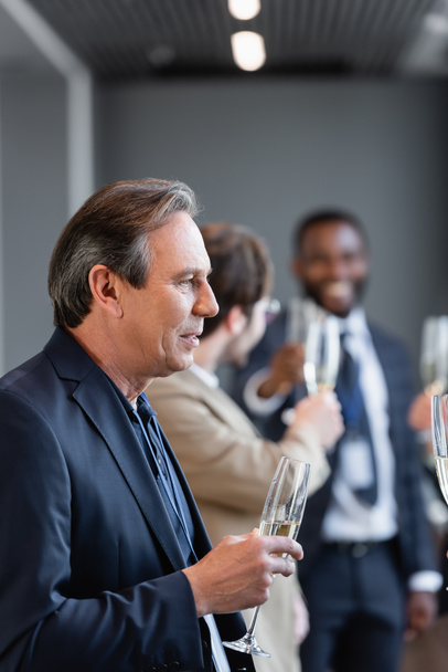 zralé podnikatel drží šampaňské sklo v blízkosti kolegů na rozmazaném pozadí - Fotografie, Obrázek
