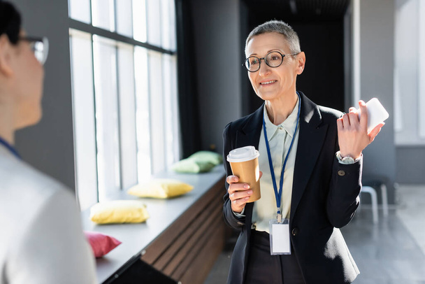 smiling businesswoman with smartphone and paper cup talking to blurred colleague - Photo, Image