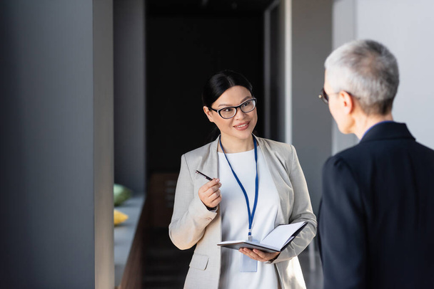 asiático mujer de negocios con notebook y pluma hablando con colega en borrosa primer plano  - Foto, Imagen