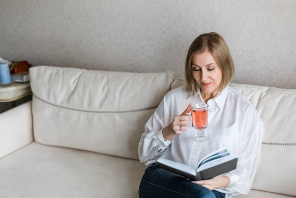 Woman is sitting on the couch holding a book in her hand and drinks tea. - Foto, imagen