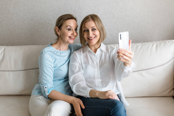 Two women sit on the couch and take a selfie on the phone while talking. - Fotó, kép