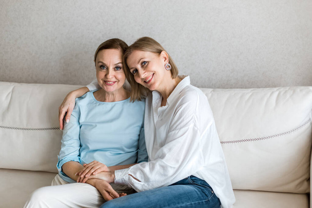 Two adult women are sitting on the couch embracing and looking at the camera. - Foto, imagen