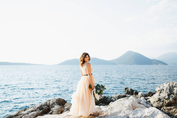 Lovely bride in a pastel wedding dress stands smiling on a rock above the sea with a bouquet of flowers in her hands - Fotoğraf, Görsel
