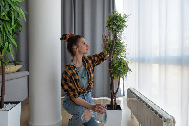 Young single woman uses her free time for the weekend and waters the flowers in flowerpot in her apartment with water - Photo, Image