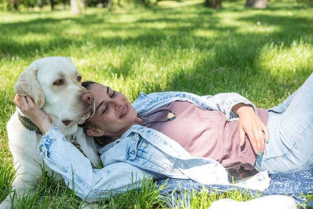 Young beautiful happy woman lying on the grass in the park holding her head on her pet dog golden Labrador Retriever and hugging him with one hand - Foto, Imagen