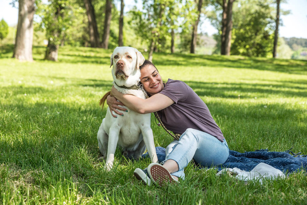 Young beautiful smiling woman sitting in the park and hugging her pet dog golden Labrador Retriever and enjoying the nice sunny weather with her friend  - Photo, Image