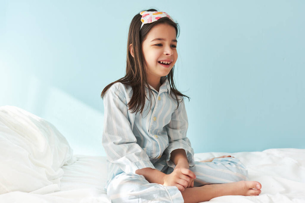 Positive little girl wearing pajama in white stripes on blue, smiling broadly, sitting in the bed after wake up in the morning, isolated on light blue background. The kid has a joyful expression. - Foto, immagini