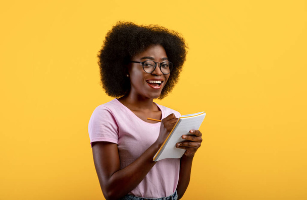Checklisten-Konzept. Schlaue schwarze Studentin mit Brille, die Notizen in einem Textbuch auf gelbem Studiohintergrund macht - Foto, Bild